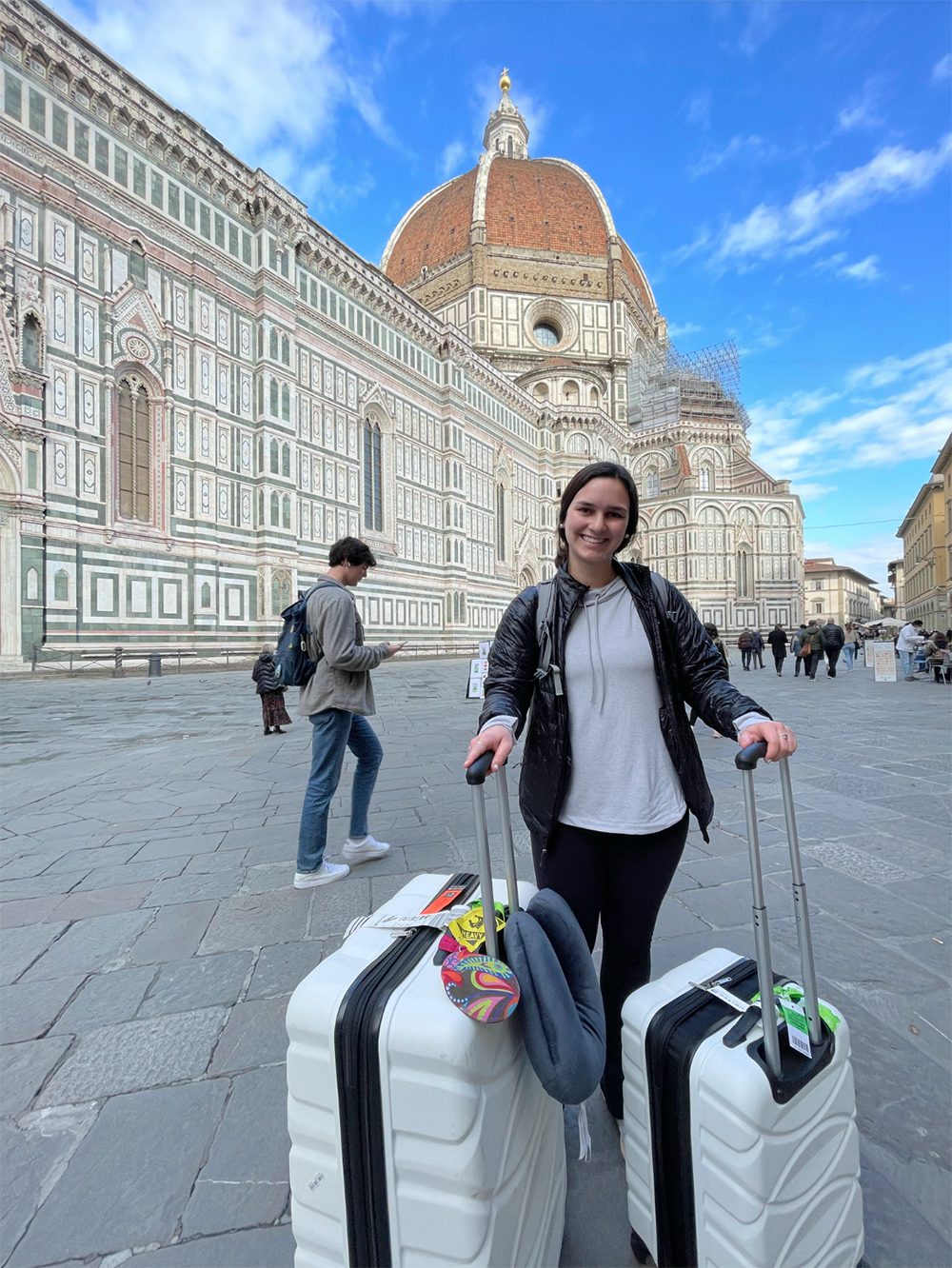 A girl wearing a white shirt ant black jacket stands in the foreground with two large white rolling suitcases, while a long white Italian building with a terra cotta dome stretching out in the background.