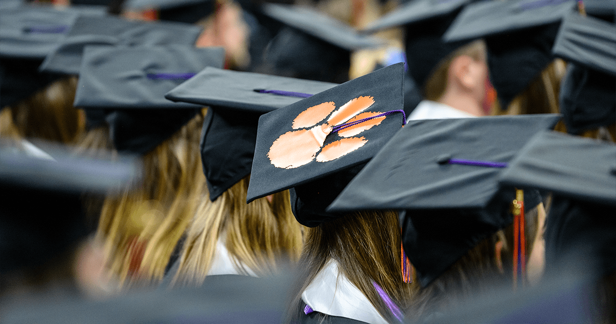 A row of students wearing graduation regalia. One cap is decorated with the Clemson University tiger paw logo.