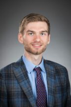 Head shot portrait of a white male with short hair and a slight beard wearing a purple tie and blue plaid blazer.