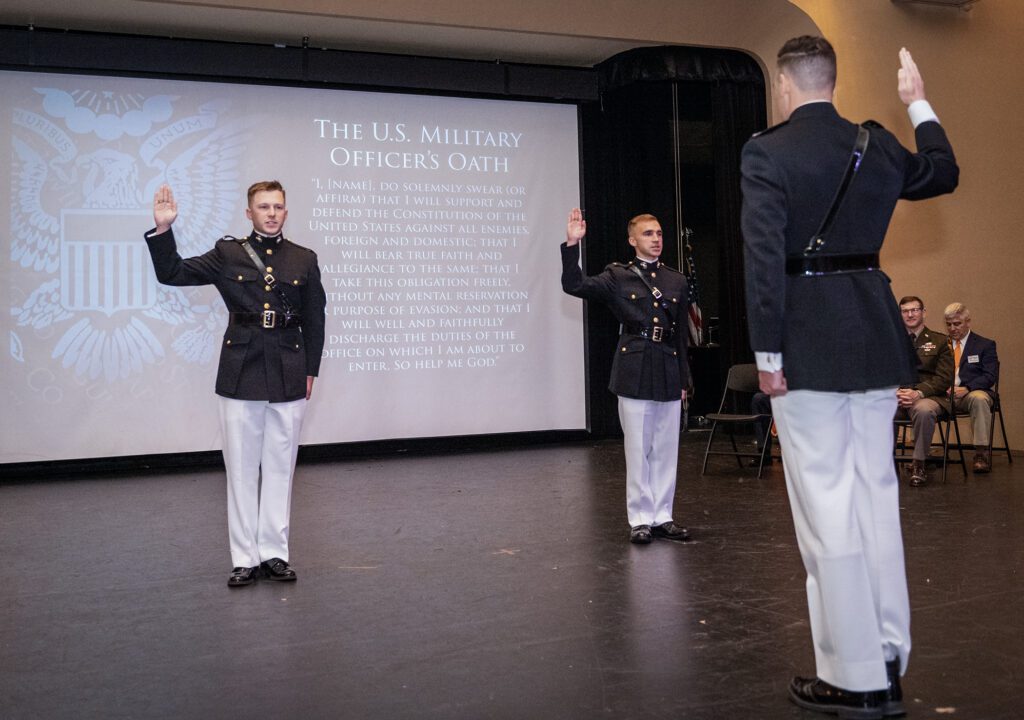 Two Marines in uniform stand at attention and hold their right hands up in front of another Marine with his back to the camera, also holding his right hand up