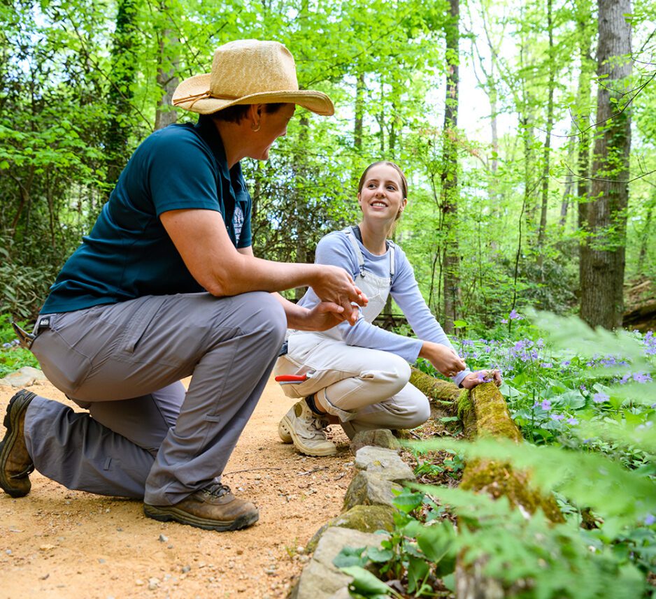 In the foreground, a woman wearing a straw hat kneels before a female student who is gardening at the edge of a planting bed. They are working in a wooded area.