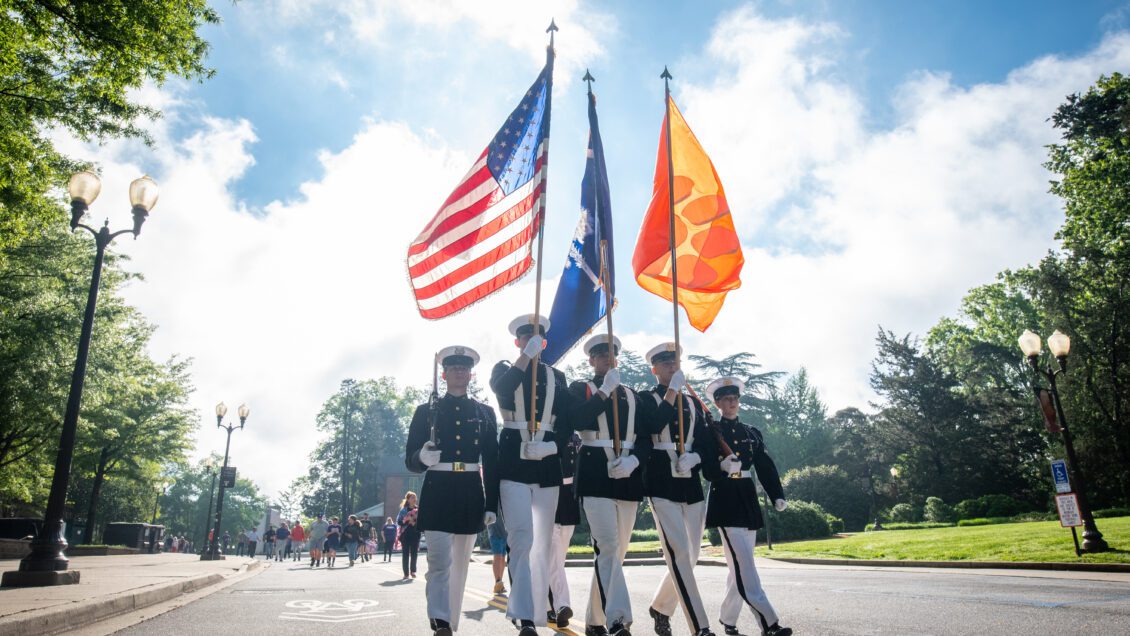 Five ROTC members walk down a road holding flag poles carrying the American flag, South Carolina flag, and Clemson flag.