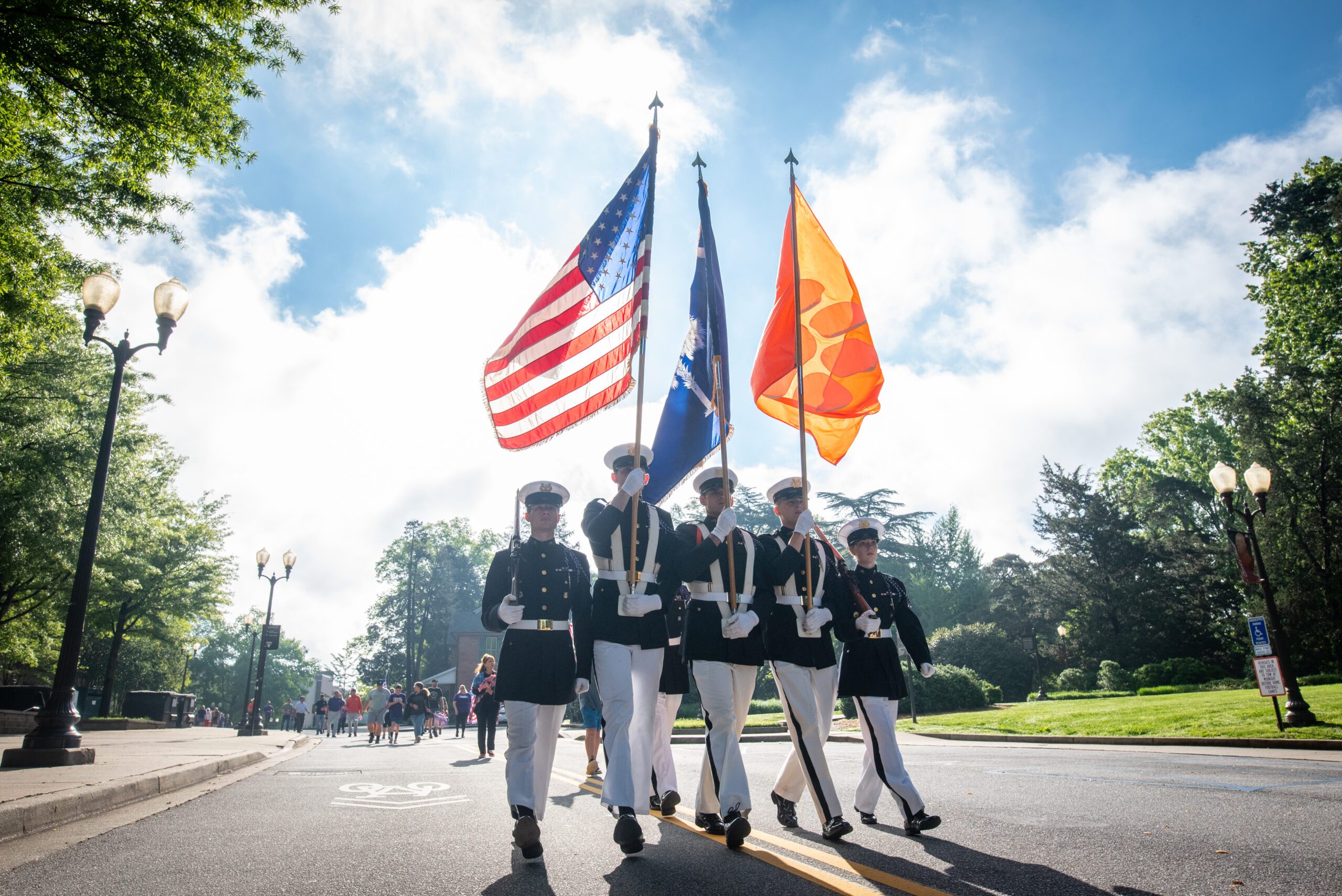 Five ROTC members walk down a road holding flag poles carrying the American flag, South Carolina flag, and Clemson flag.