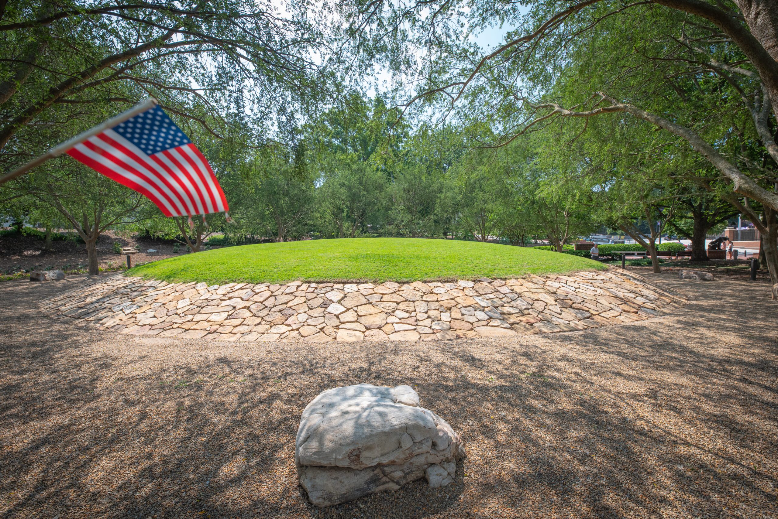 An American flag blows in the upper left corner of the frame in front of a mound ringed by rocks and topped by thick green glass, immaculately manicured, sits amid rows of trees.