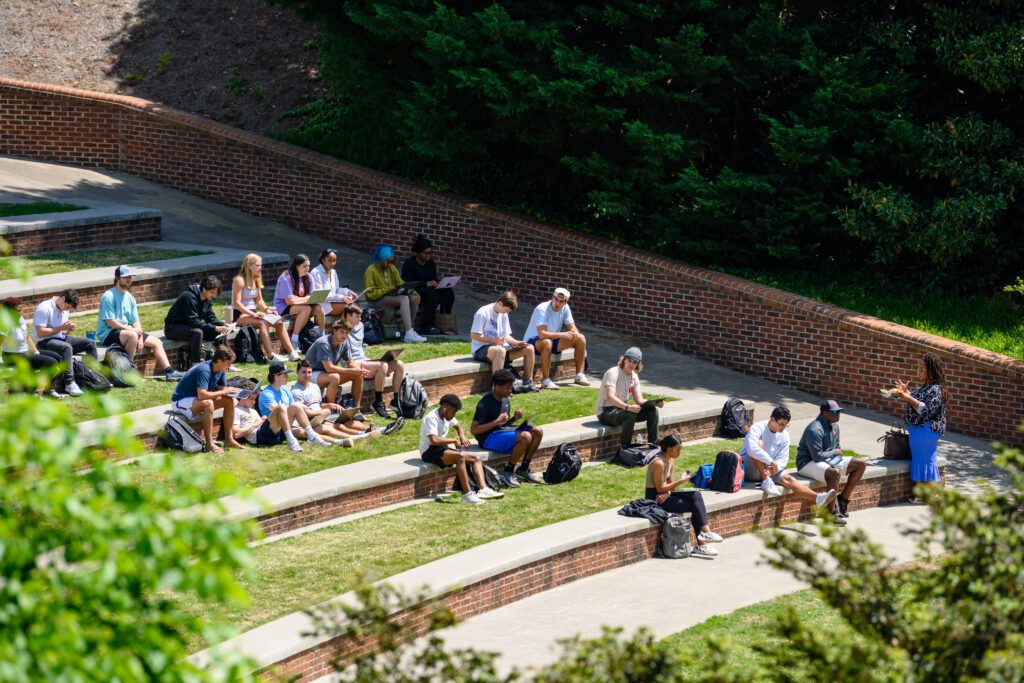 An outdoor Spring semester class with a diverse group of students sitting on amphitheater style seating of brick and concrete bleachers and grass layers, and the professor standing at the bottom of the hill. The sun is shining, however a thick grouping of trees and shrubs provide some shade for students near the top of the hill.