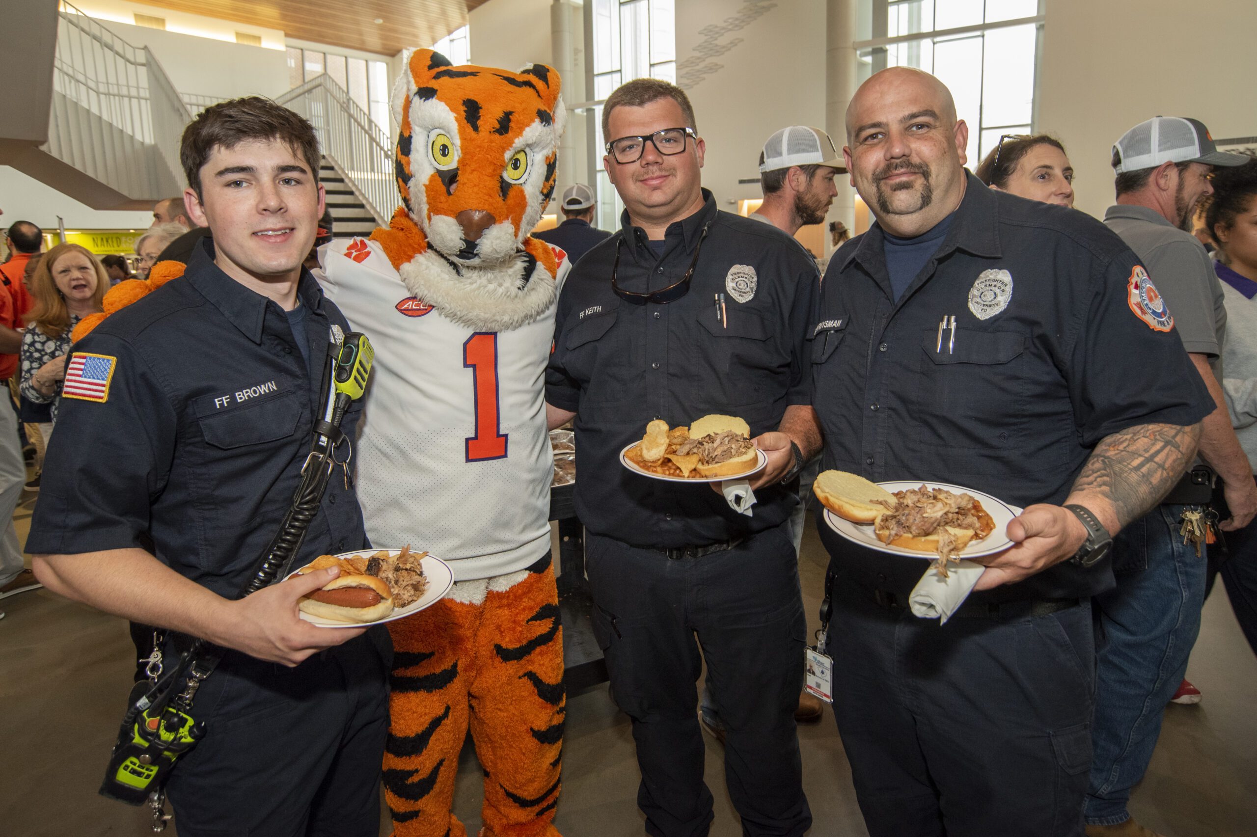 Three men wearing Clemson Police uniforms and holding plates of food pose for a photo with the Clemson Tiger.