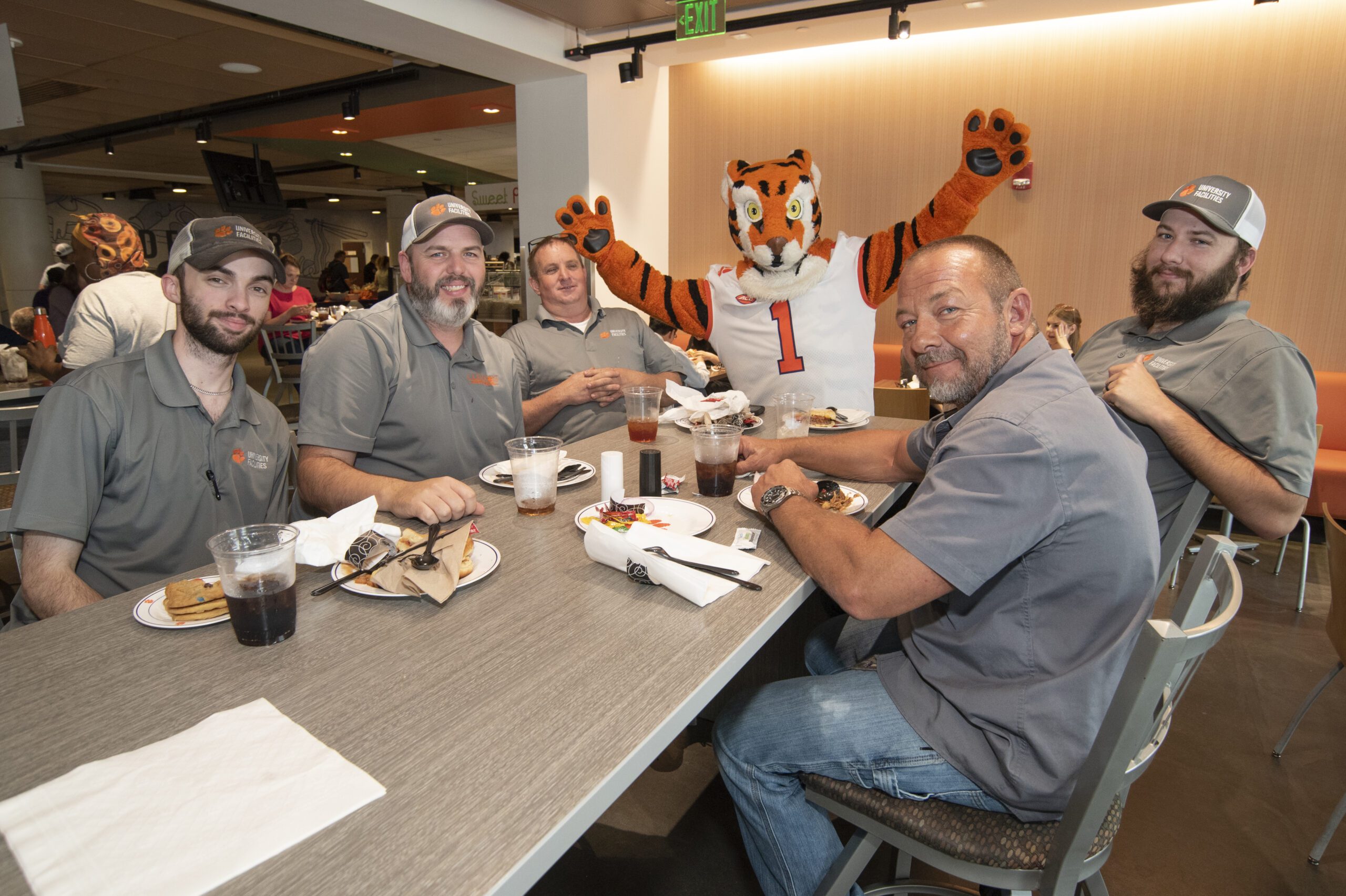 Five men dressed in grey polos sit at a table around the Clemson Tiger mascot while smiling for a photo.