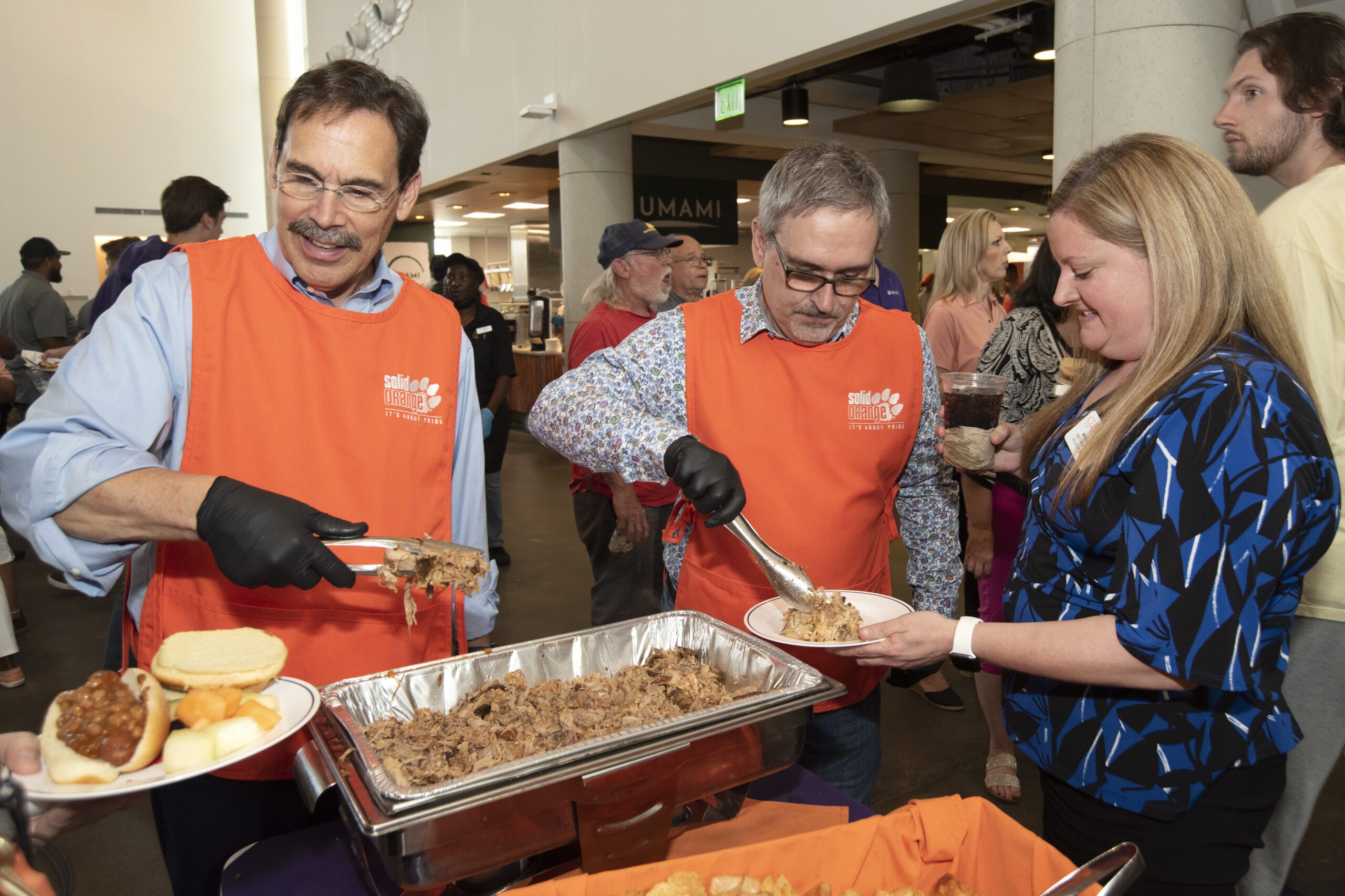 Two men wearing glasses, button up shirts, and Solid Orange aprons serve food to a woman in a blue shirt.