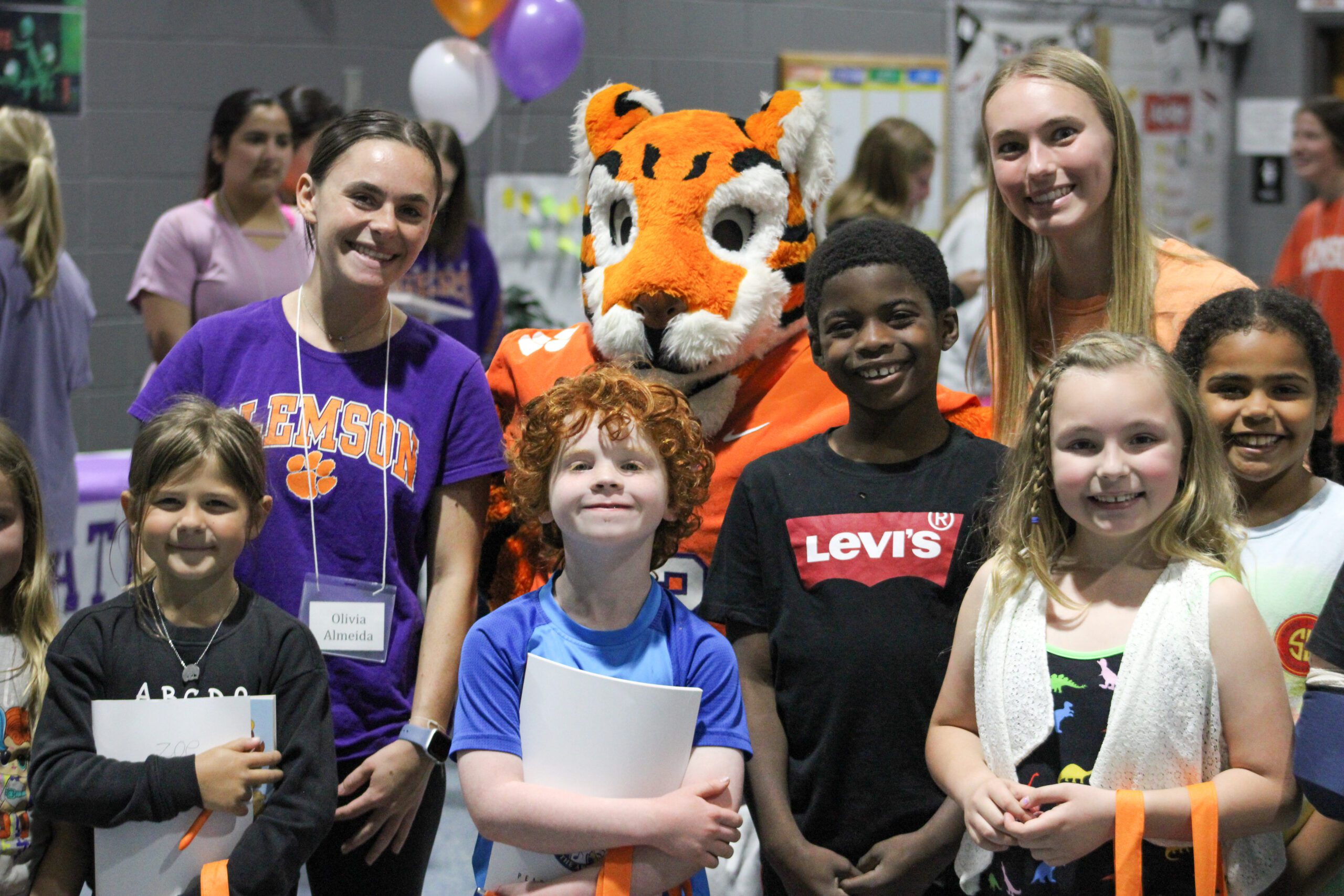 Students from Clemson University's Pearce Center for Professional Communication pose for a photo with elementary students at Central Academy of the Arts and the Clemson Tiger Cub mascot. The children are holding books.