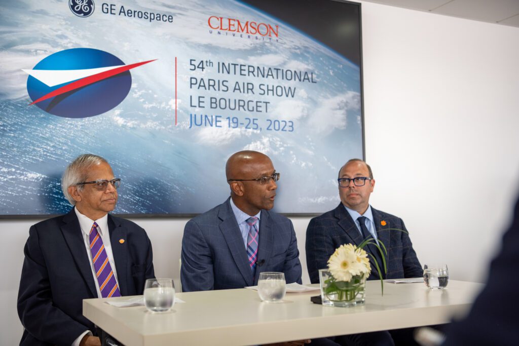 Three men wearing suits are seated in front of a Paris Air Show GE Aerospace and Clemson University banner