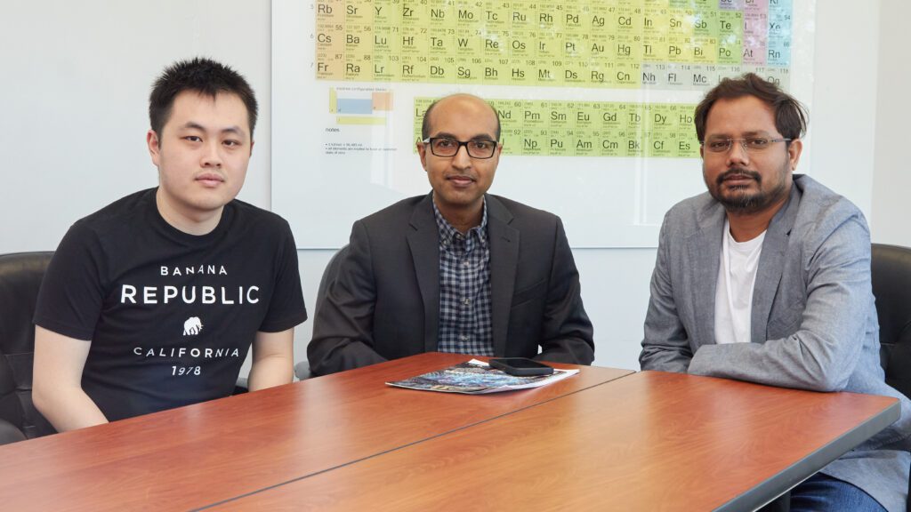 Three men sitting at a table in front of a poster of the periodic table