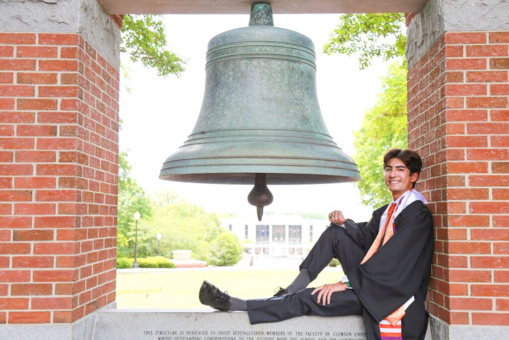 Gabe Cutter, wearing graduation robes, sitting by the Old Tillman Hall Bell in the Carillon Gardens on Clemson University campus. 