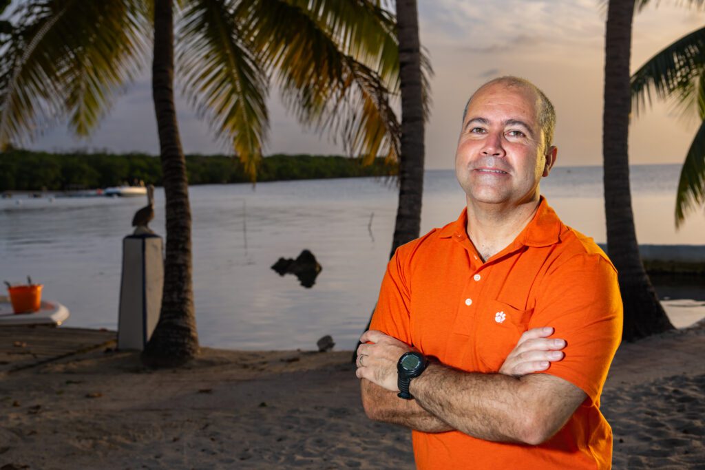 A Latino man wears an orange shirt and is standing with his arms crossed in front of a palm tree and waterfront at dusk.