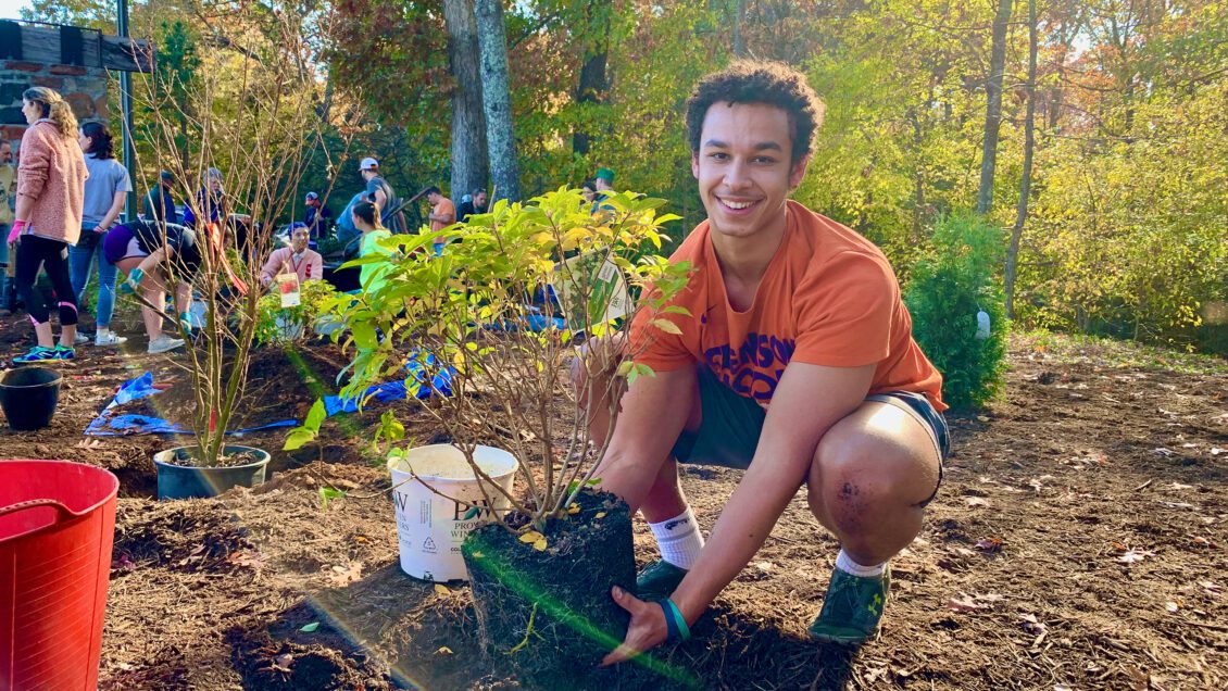A young male, a student of the new STEM designated landscape architecture student poses in front of the camera with a smile on his face. He is squatting down and holding a small tree in his hands that it looks like he is getting ready to plant.