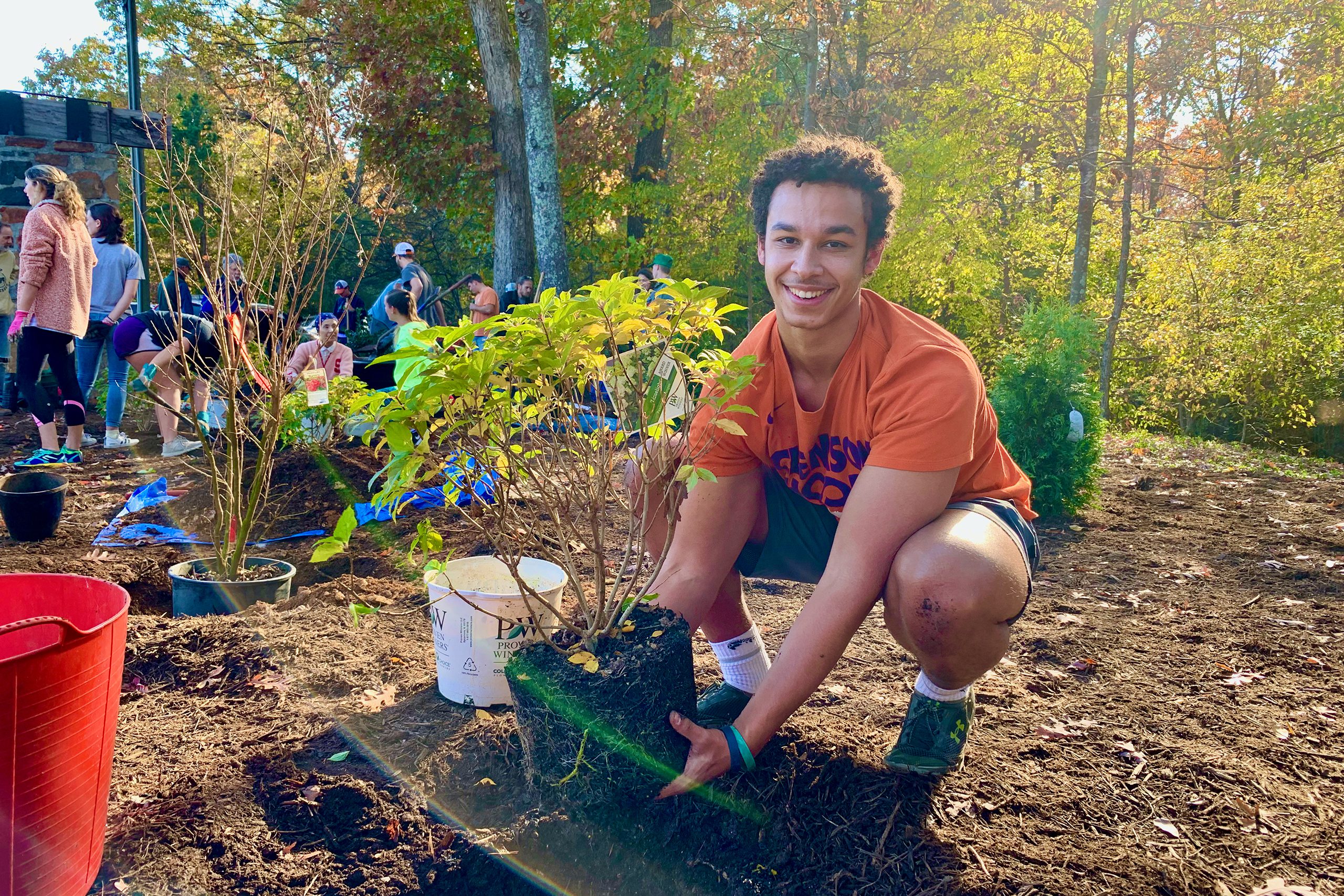 A young male, a student of the new STEM designated landscape architecture student poses in front of the camera with a smile on his face. He is squatting down and holding a small tree in his hands that it looks like he is getting ready to plant.