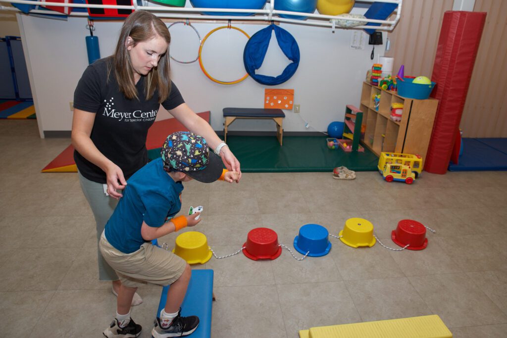 A woman wearing a black shirt helps a boy walk over a step during a physical therapy session.