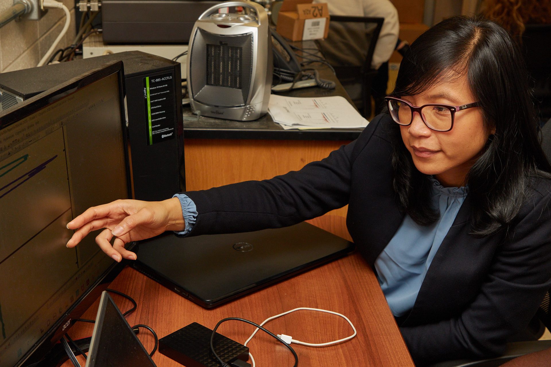 Woman wearing glasses and a black suit points at a computer screen.