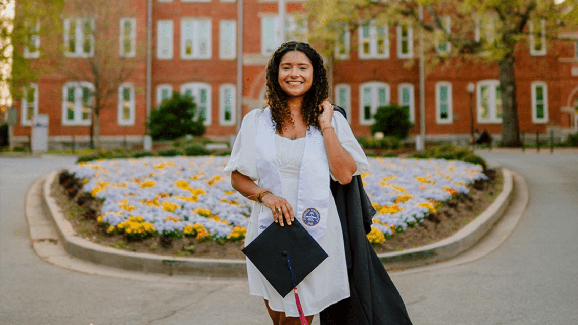 Portrait of Aracely Quintero with graduation cap in front of a bed of colorful flowers and a brick academic building