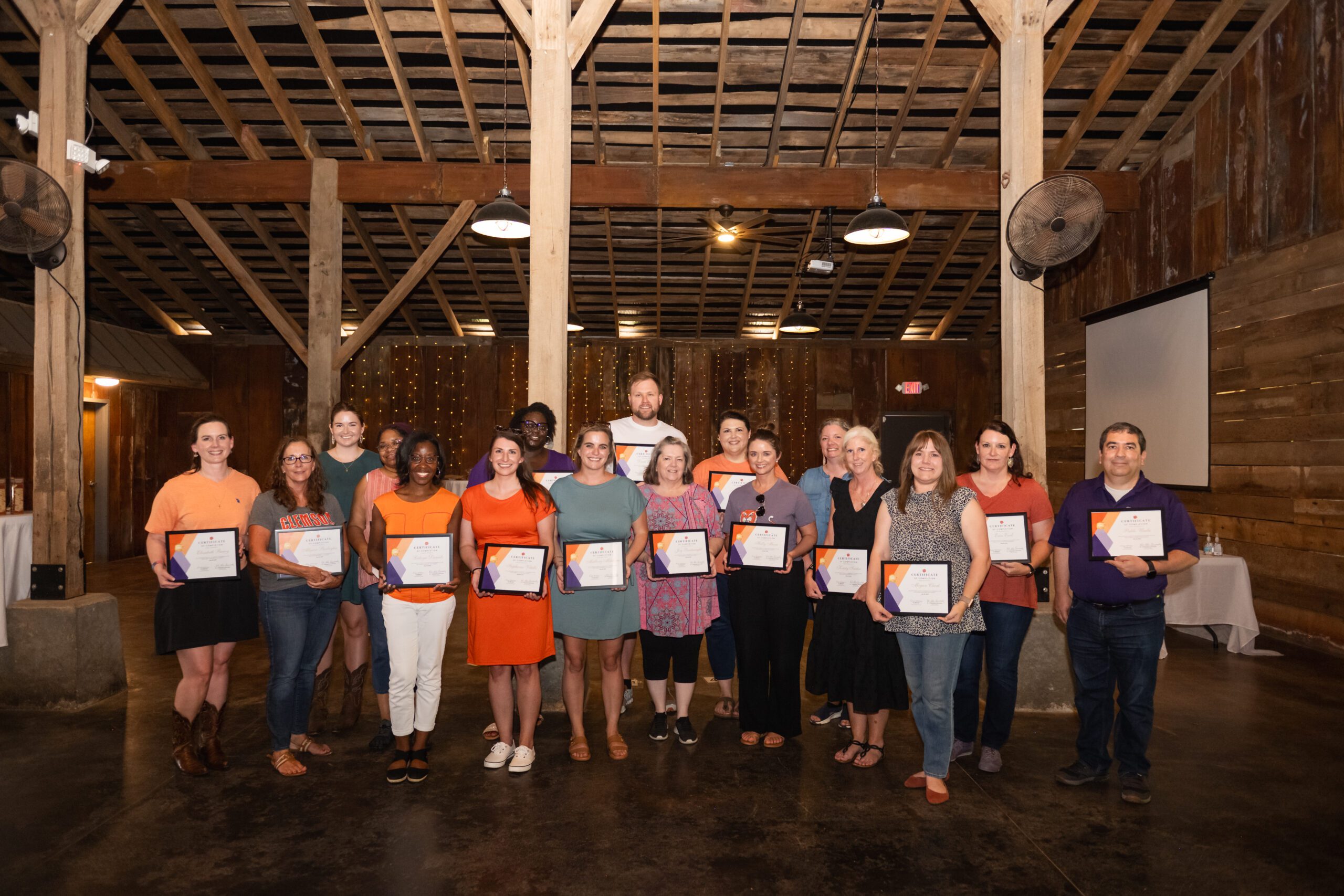 17 people stand in a group holding certificates inside a barn with tall wooden columns.