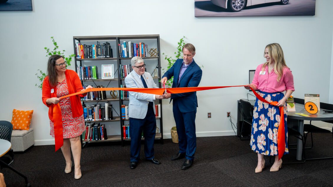 Two women hold a large orange ribbon while two men in the center cut the ribbon. There is a bookshelf in the background.