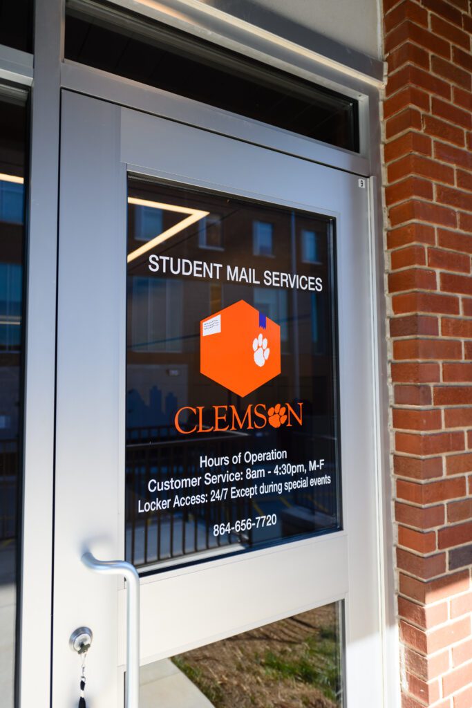 The front door of the student post office. The door reads "STUDENT MAIL SERVICES" with customer service hours of operation 8 am-4:30 p.m., M-F, and locker access 24/7 except for special events. 864-656-7720