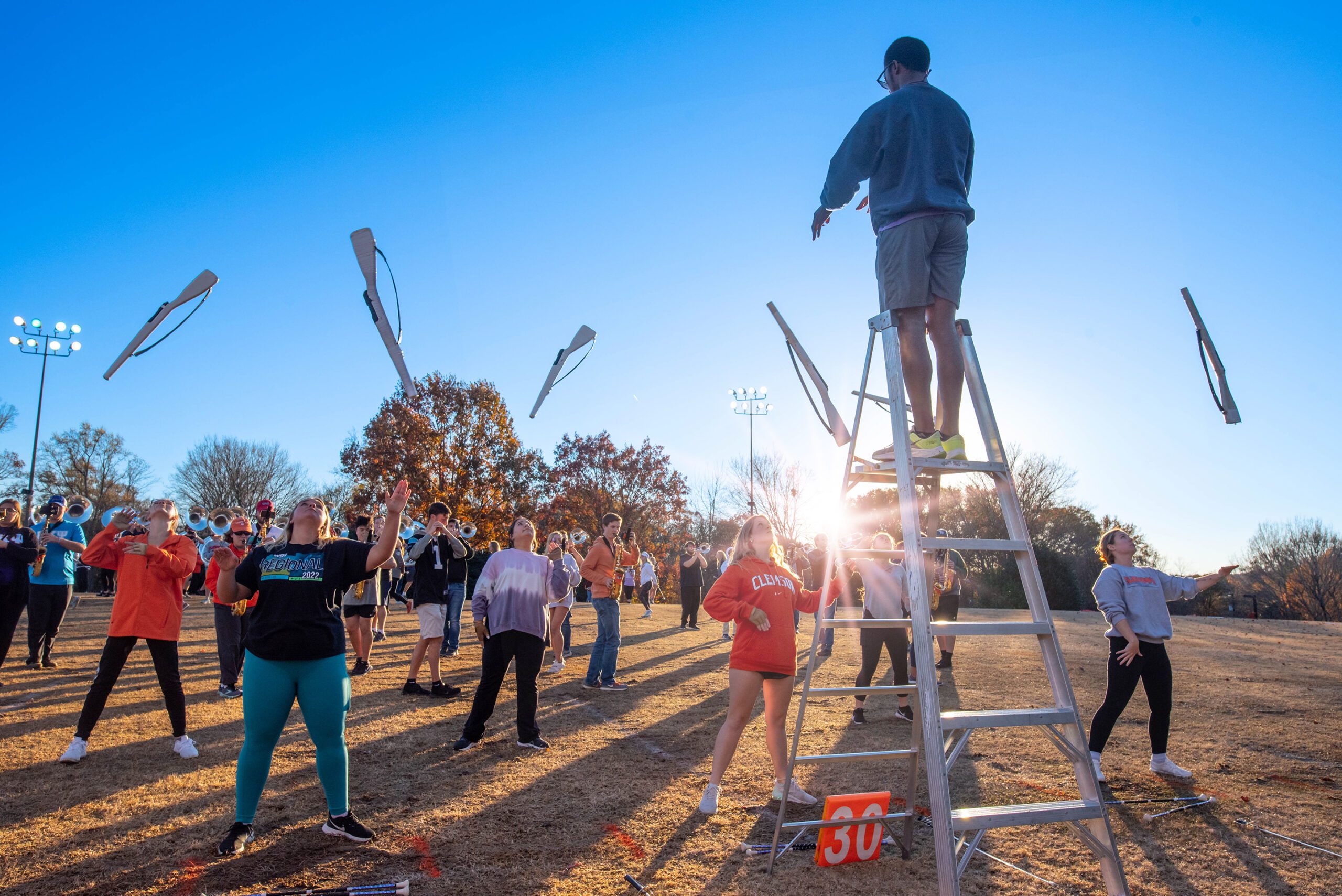 A man on a ladder directs a row of students tossing rifles into the air