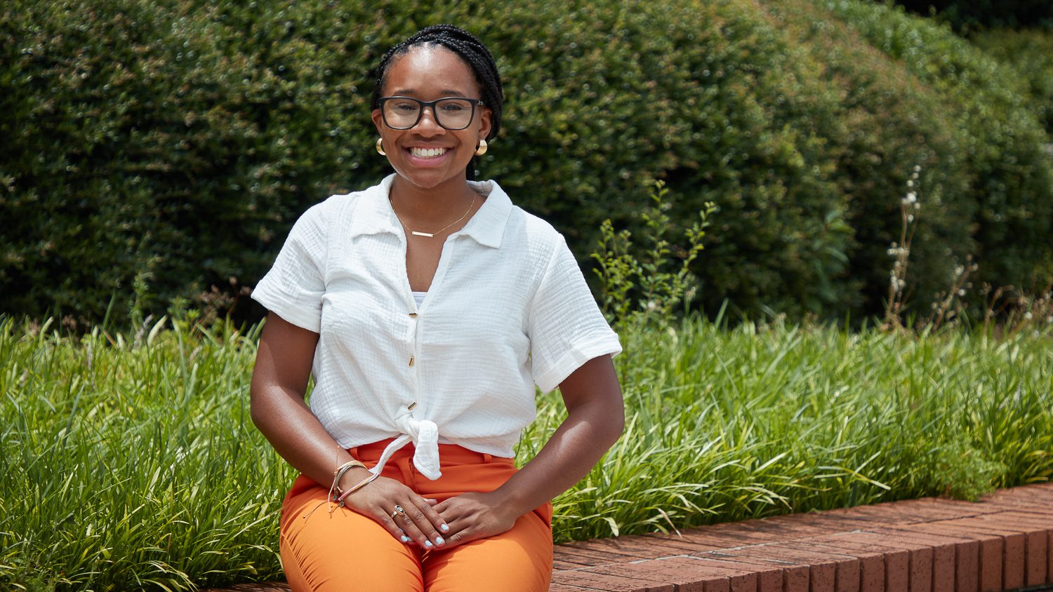 Black woman wearing a white shirt and orange pants sits on a brick ledge in front of greenery.