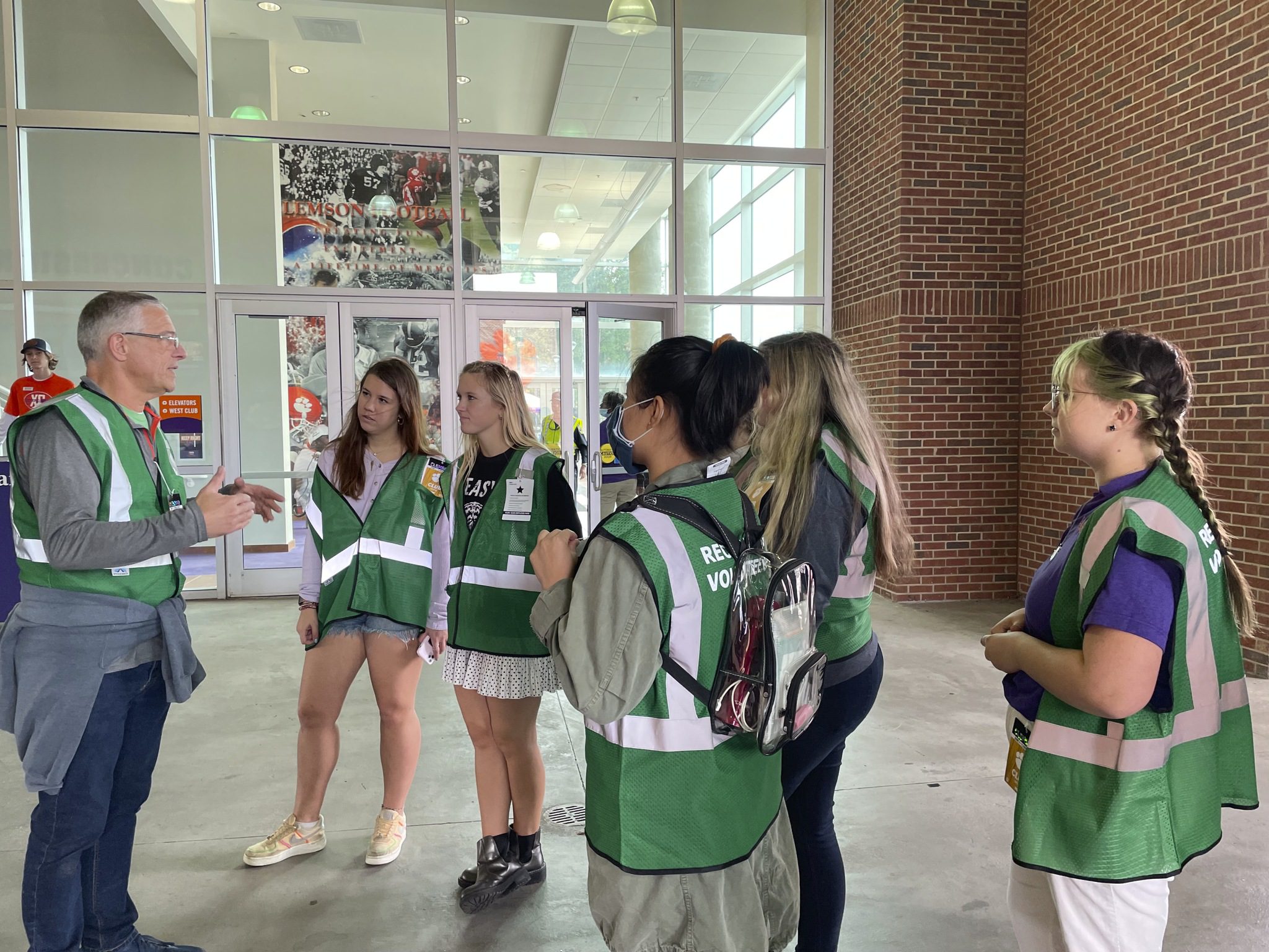 Five women in a green reflective pull over talk with a man in a green reflective pullover.