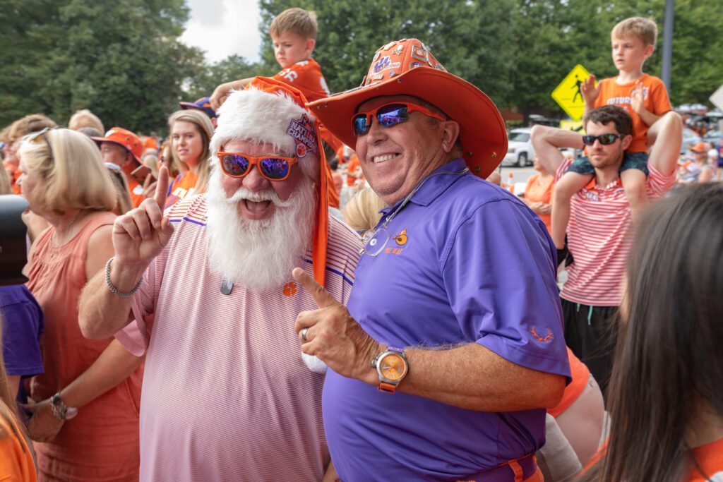 Two Clemson fans, one dressed as Santa Claus and one with an orange cowboy hat wear sunglasses and put their index fingers in the air. 