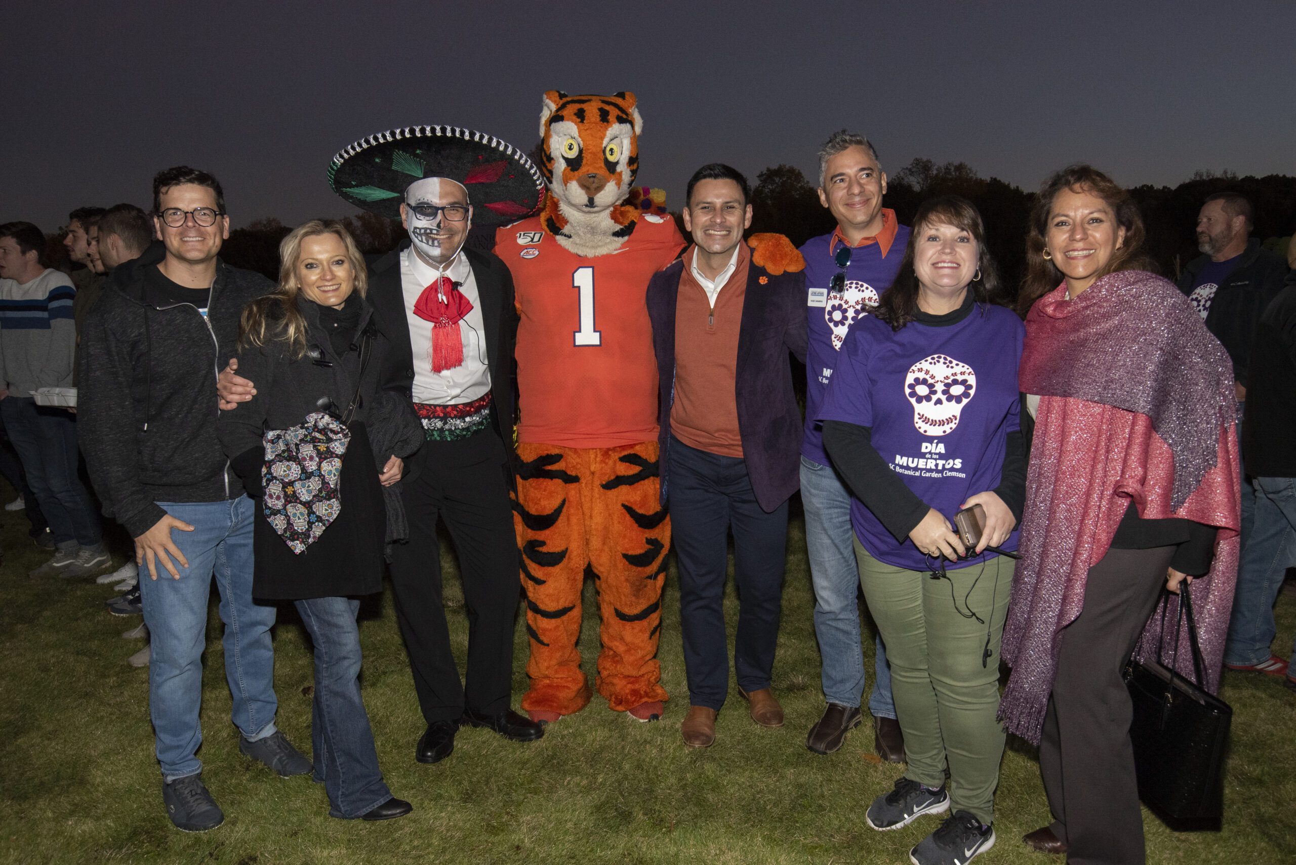 A nighttime image of a group of 6 people dressed in orange and purple and one dressed in a skeleton Day of the Dead suit with a sombrero gather around the Clemson Tiger cub.