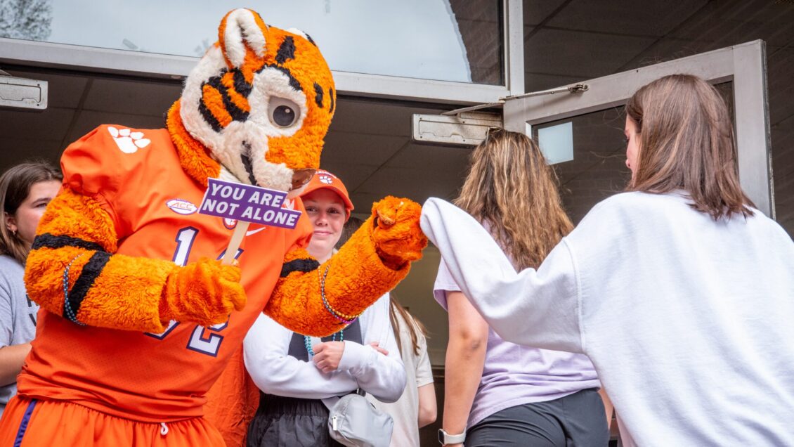 The Clemson Tiger fist-pumping with a student while holding a sign that says "You Are Not Alone"