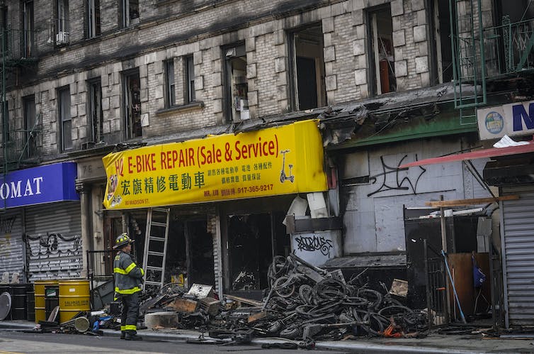 A firefighter stands outside a charred stone building. A pile of burned bicycles  sits under a sign reading "E-Bike Repair (Sale and Service)".