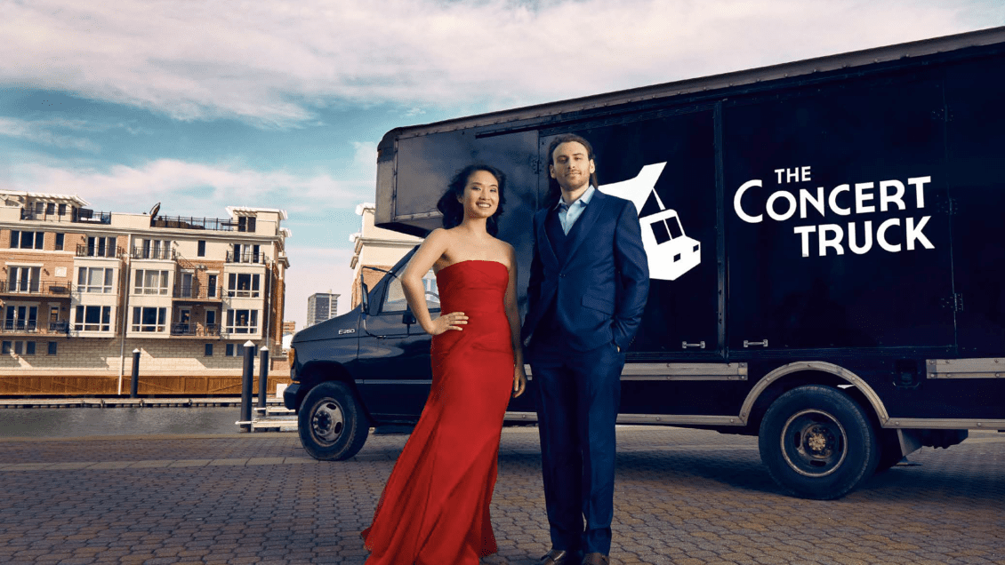 A woman in a red designer dress and a man in a tux stand in front of a 16' truck with the words The Concert Truck labeled on its sides.