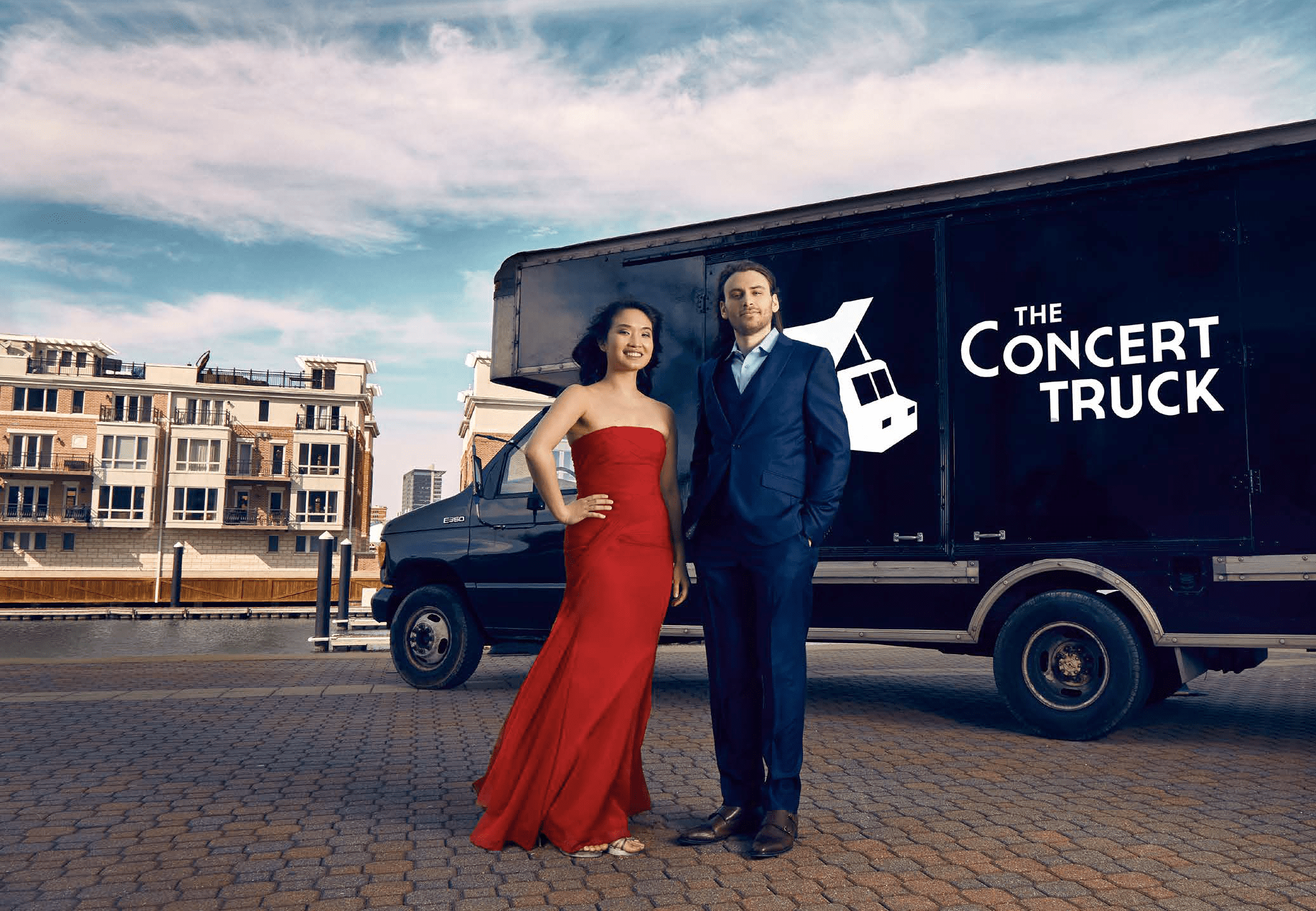 A woman in a red designer dress and a man in a tux stand in front of a 16' truck with the words The Concert Truck labeled on its sides.
