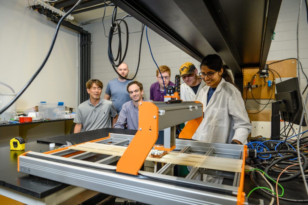 A group of researchers gather around a large, table-like orange and grey device in a lab. 