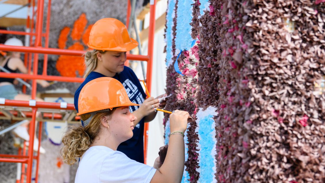 Two young women waring orange hardhats are working with tissue paper on a large homecoming. Orange scaffolding is in the background.