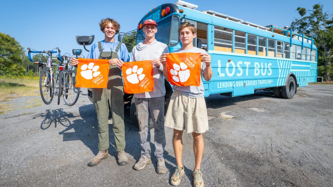 Three young men stand in front of a bus holding Clemson Tiger Rags