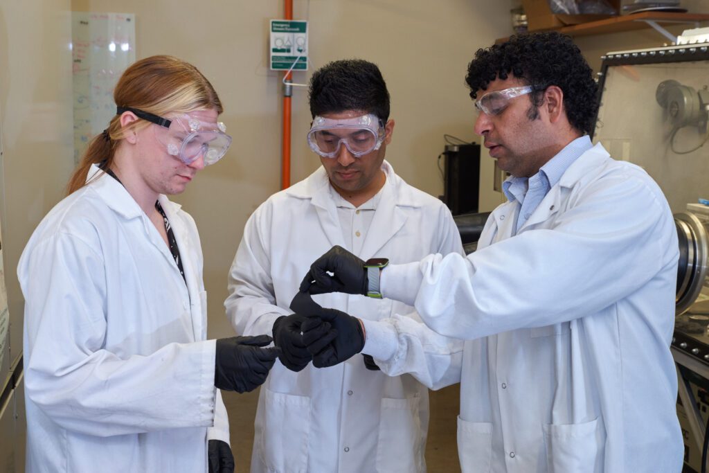 three men in white lab coats looking at a square piece of metal.