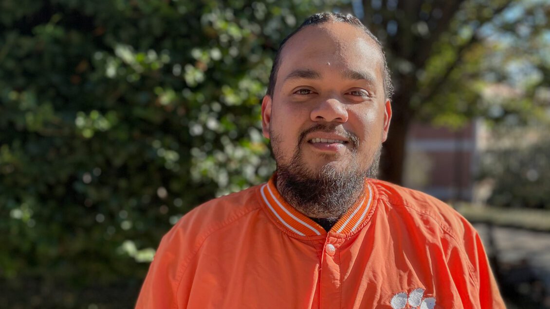 Melvin Villaver is pictured in an orange Clemson Tigers Starter Jacket near the Carillon Garden on a sunny fall afternoon.