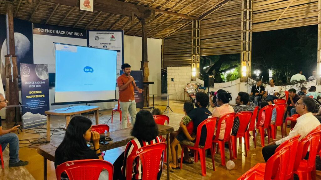 a man wearing an orange shirt makes a presentation in front of an audience.