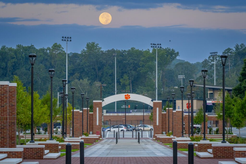 A photo of a long concourse at early morning features a tiger paw on a concrete arch and a full moon against a blue backdrop of sky is overhead. 