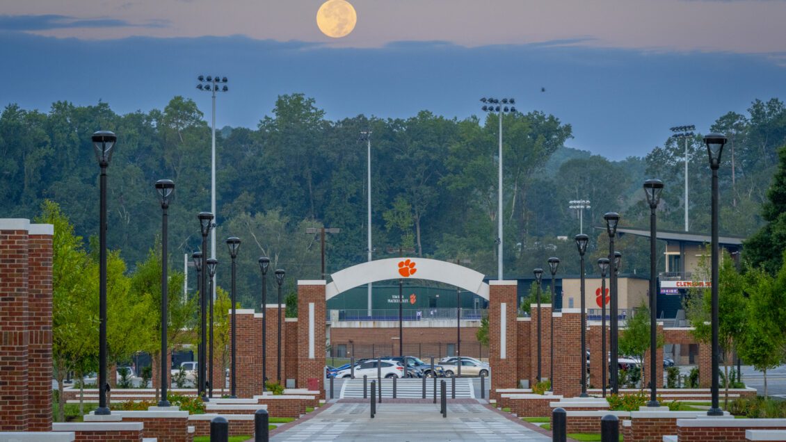 A photo of a long concourse at early morning features a tiger paw on a concrete arch and a full moon against a blue backdrop of sky is overhead.