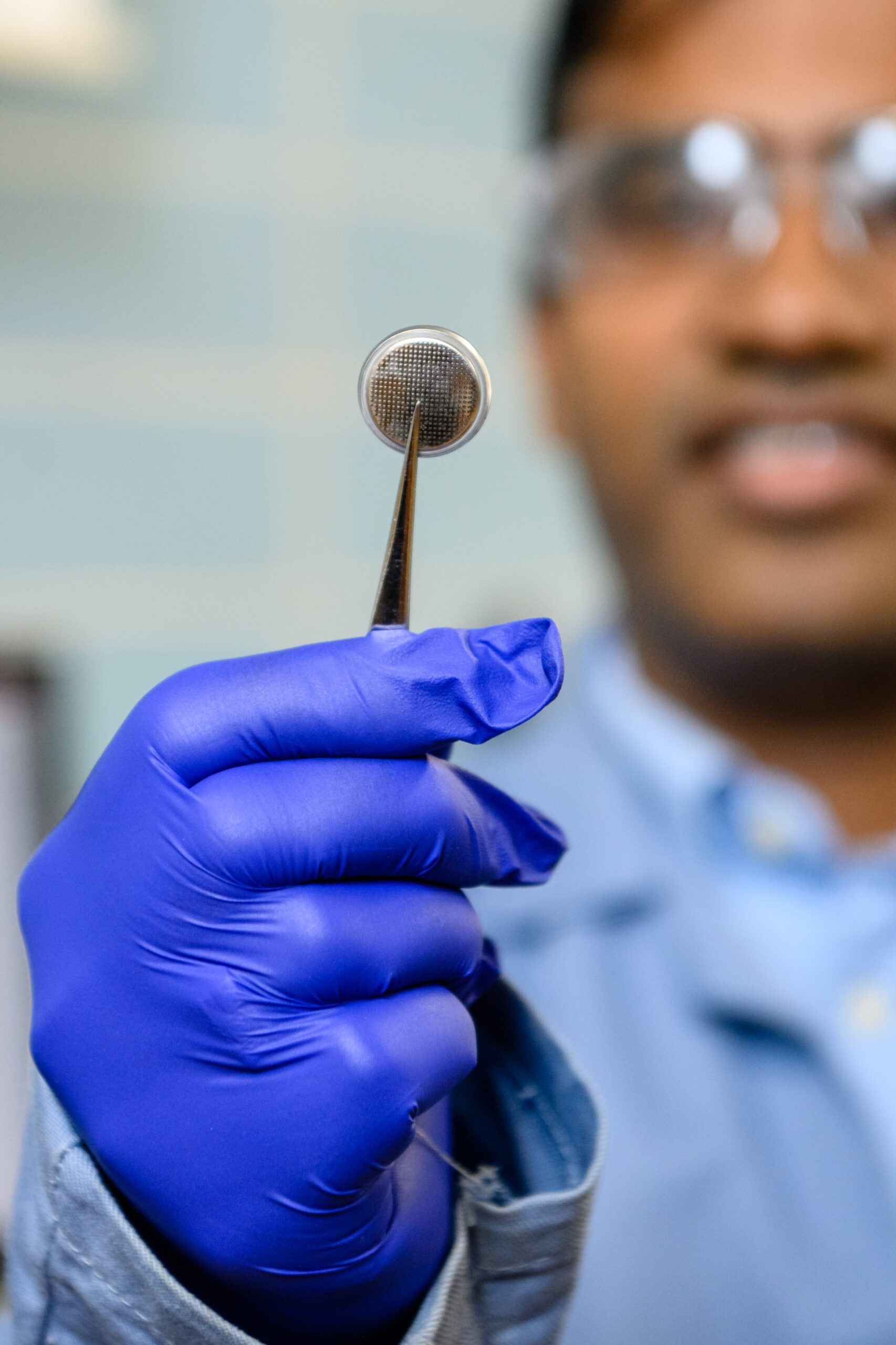 In the foreground focus is a blue-gloved hand holding a set of tweezers that are pinching a circular metal and mesh looking battery with face of a man wearing safety goggles in the background. 