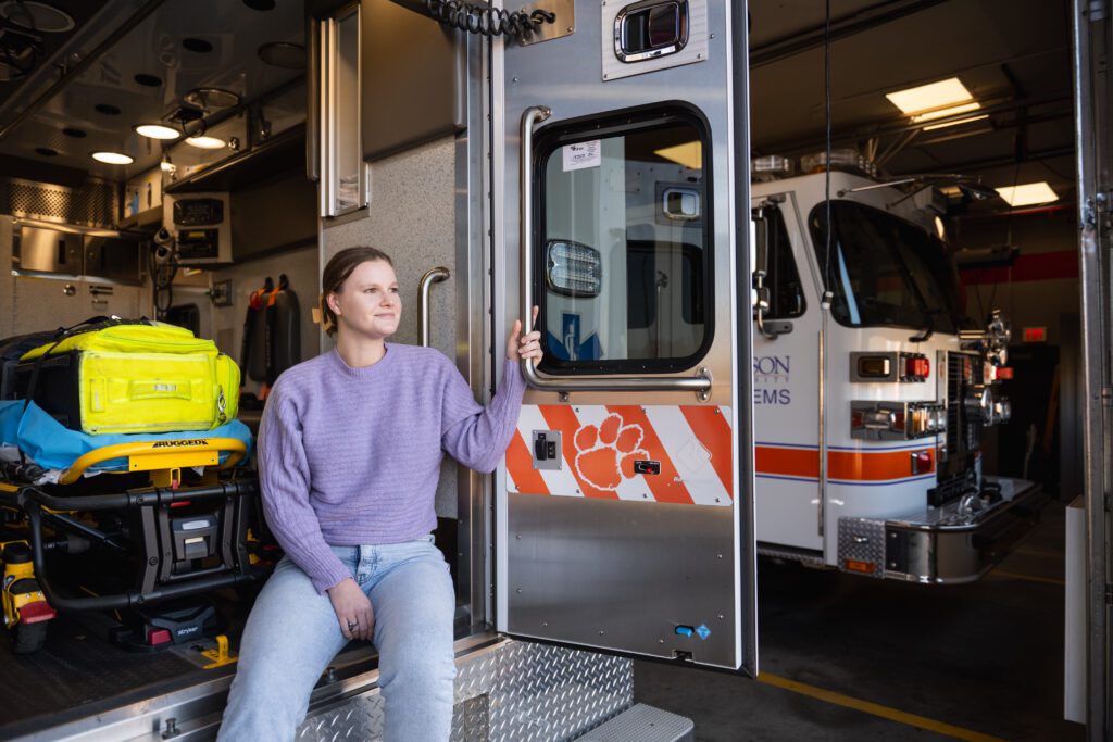 A young woman with her hair tied back sits on the back of an ambulance that is parked in a bay. The back door is open and it shows a Clemson Tiger Paw on the door and medical equipment inside. 