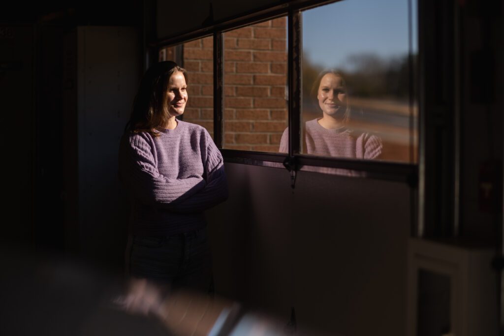 A young woman wearing a long-sleeve purple shirt sits at a window and is looking out on a view of the Clemson campus.