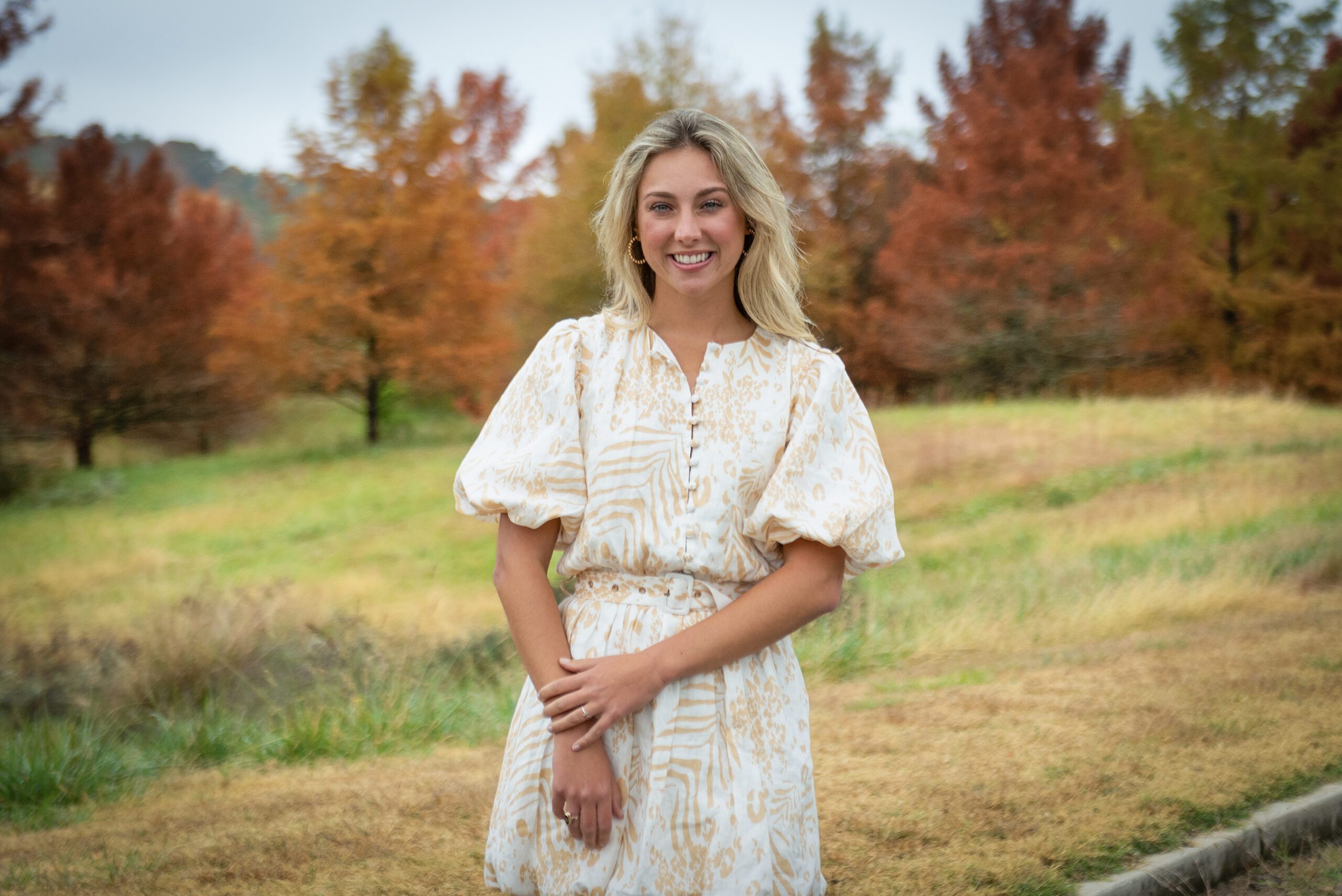 A young lady in a dress stands in a green field with trees full of colorful fall leaves behind her