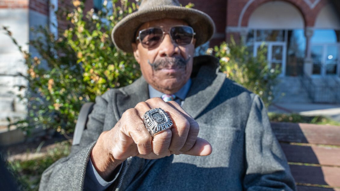 An older gentlemen dressed in a dapper coat, fedora and sunglasses holds his hand out to the camera to show off a large ring.
