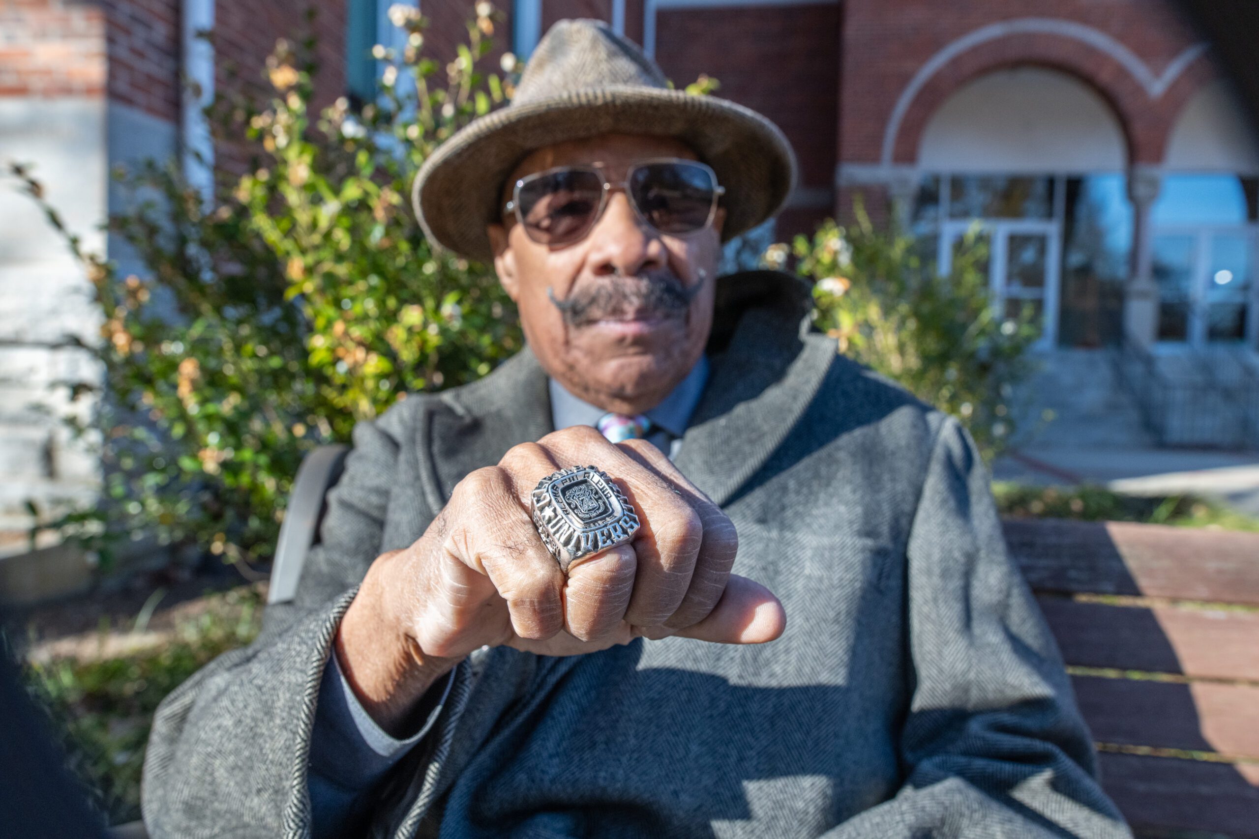 An older gentlemen dressed in a dapper coat, fedora and sunglasses holds his hand out to the camera to show off a large ring.