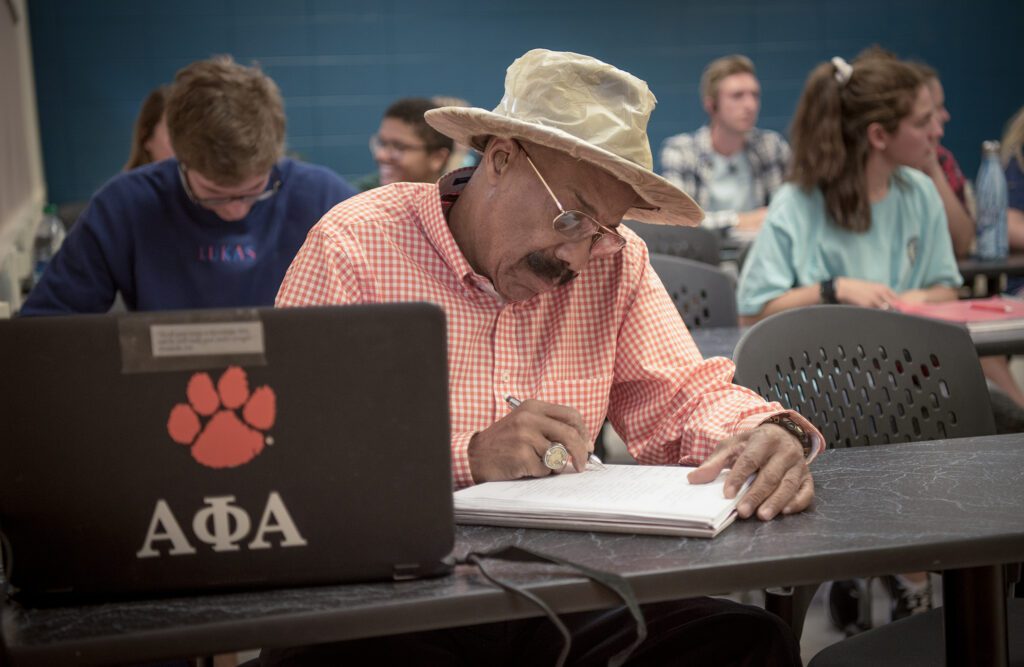 An older gentleman wearing a fedora sits at a school desk taking notes