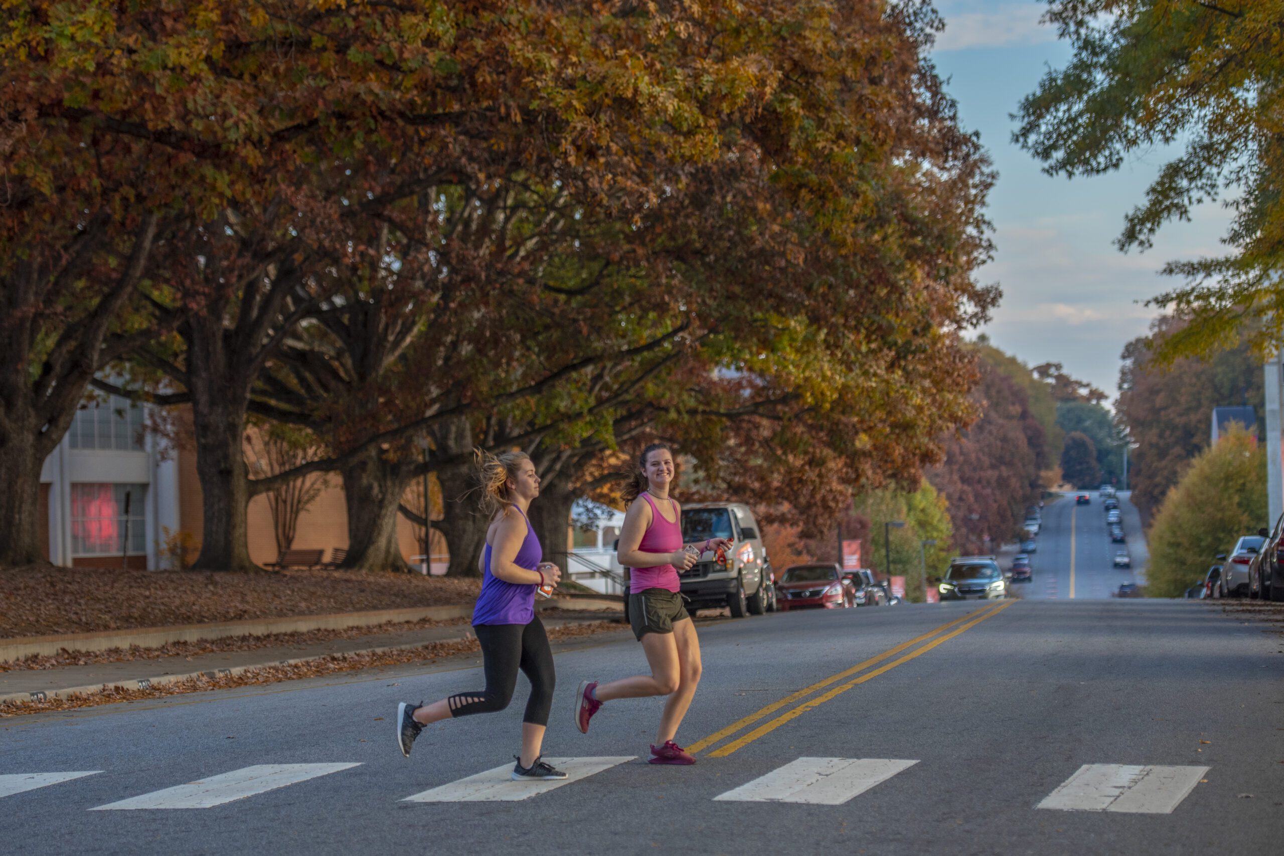 Two female runners jog over a crosswalk with fall trees and lit buildings behind them, indicating morning.